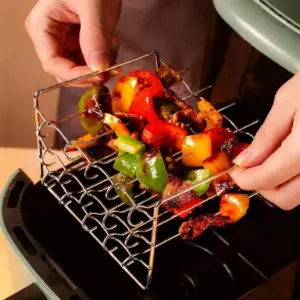 Hand hält ein Metallgestell mit Paprika und Fleischstücken über einem Airfryer