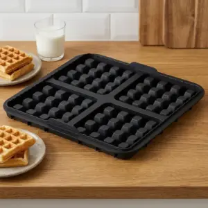 Schwarze Waffelform mit quadratischen Mulden auf einem Holztisch, daneben Waffeln